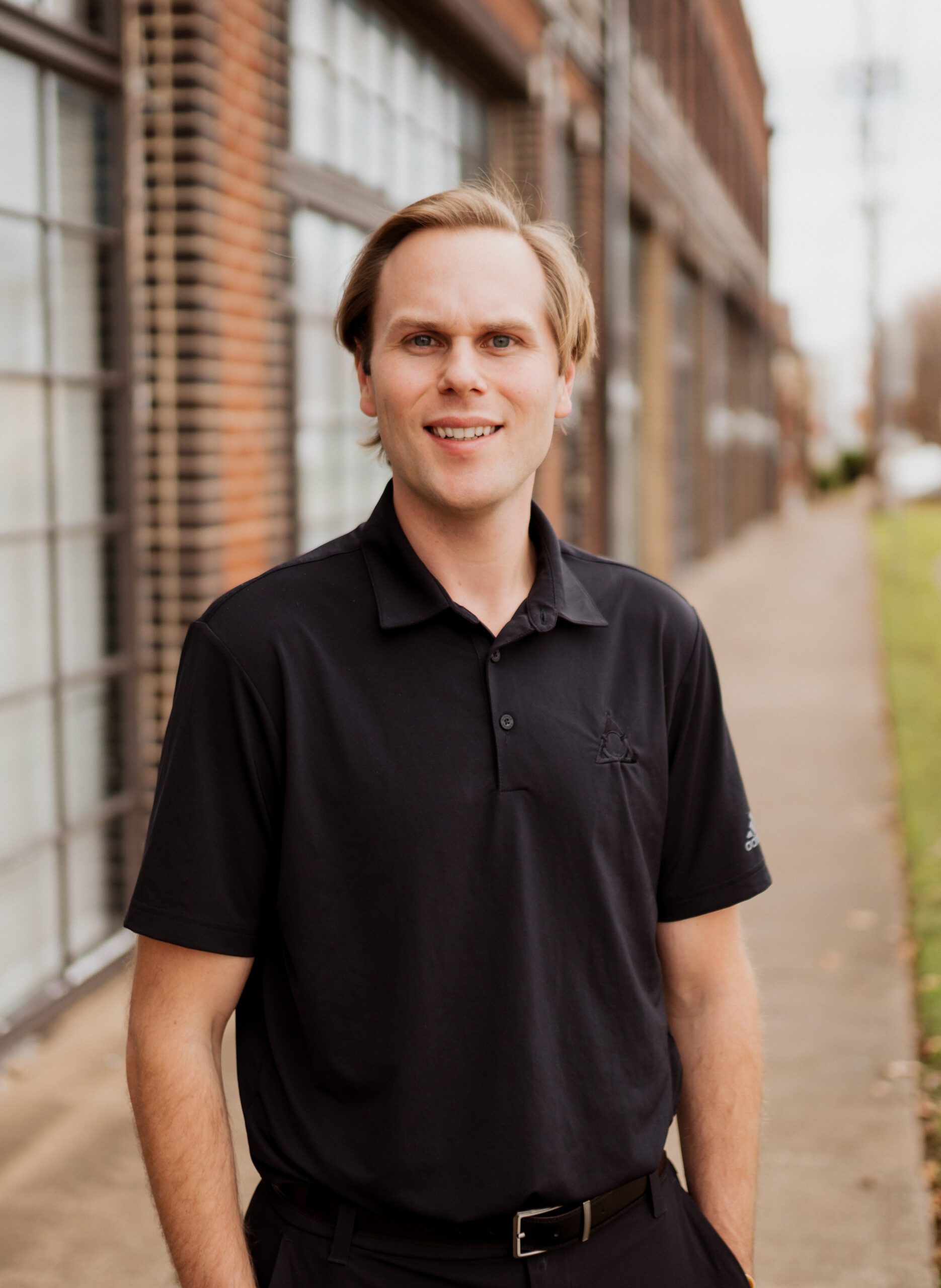 Michael Dunham smiling with hands in his pockets standing in front of a brick wall wearing a black polo shirt